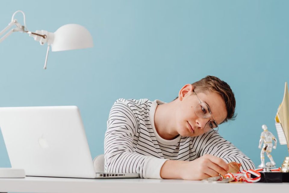 Teenager with eyeglasses contemplating medals at desk with laptop and trophies.